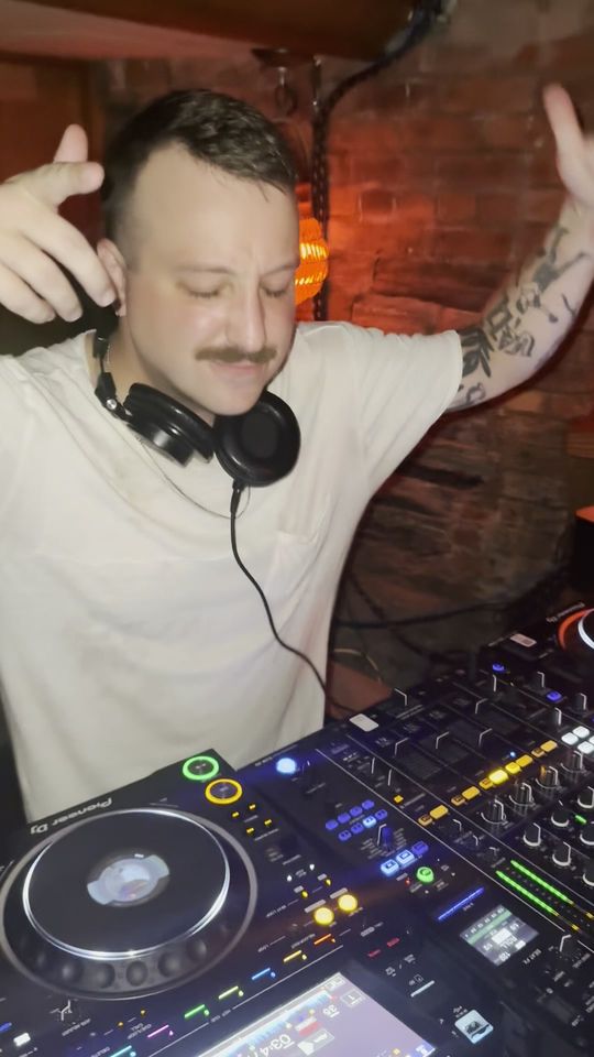 DapSoul in a white T-shirt, both arms up behind the decks